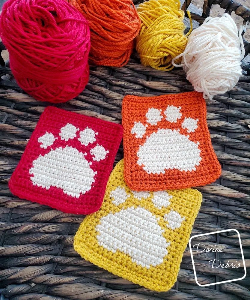 [Image description] A slightly angled view of 3 colored Cute Paw Print Squares, one orange, one yellow, and one red, in a wooden woven basket with balls of yarn along the top of the basket. 