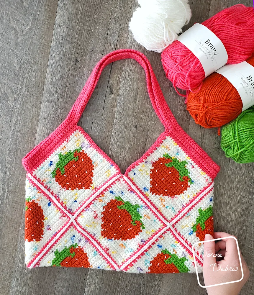 [Image description] The Cute Strawberry Bag laying flat on a fake wood grain background with skeins of yarn along the top right of the photo and a white woman's hand holding the bottom right corner of the bag.