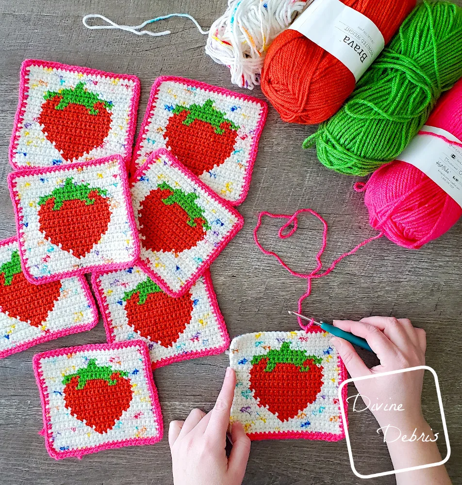 [Image description] Top down view of the a bunch of Cute Strawberry squares and a white woman's hands finishing a border of a square on a wood grain background.