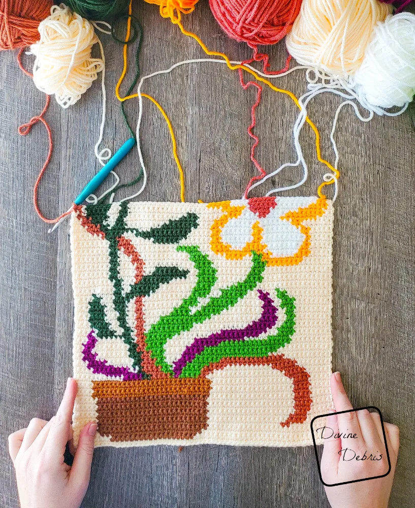 [Image description] Top down view of the unfinished Groovy Bloom Wall Hanging with a white woman's hands holding the bottom 2 corners and skeins of yarn attached to the hanging at the top.