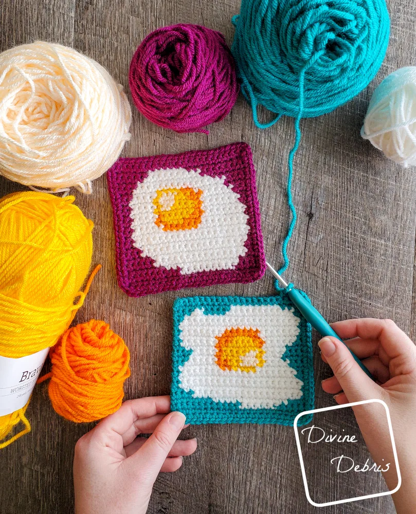 [Image description] Top down view of the Sunny Side Up Egg Squares, laying on a fake wood background with skeins of yarn along the top and left side of the photo and a white woman's hands crocheting a border on the blue square.