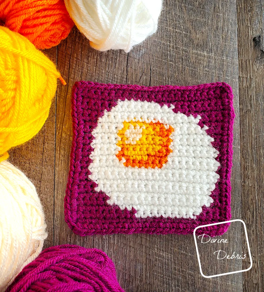 [Image description] Top down view of the Sunny Side Up Egg Square 1, laying on a fake wood background with skeins of yarn along the left side of the photo.