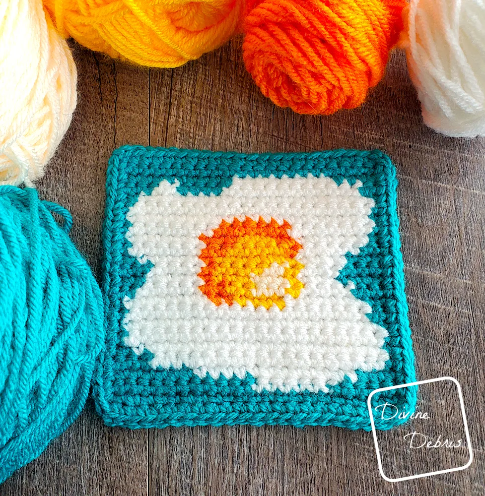 [Image description] Top down view of the Sunny Side Up Egg Square 2, laying on a fake wood background with skeins of yarn along the top of the photo.