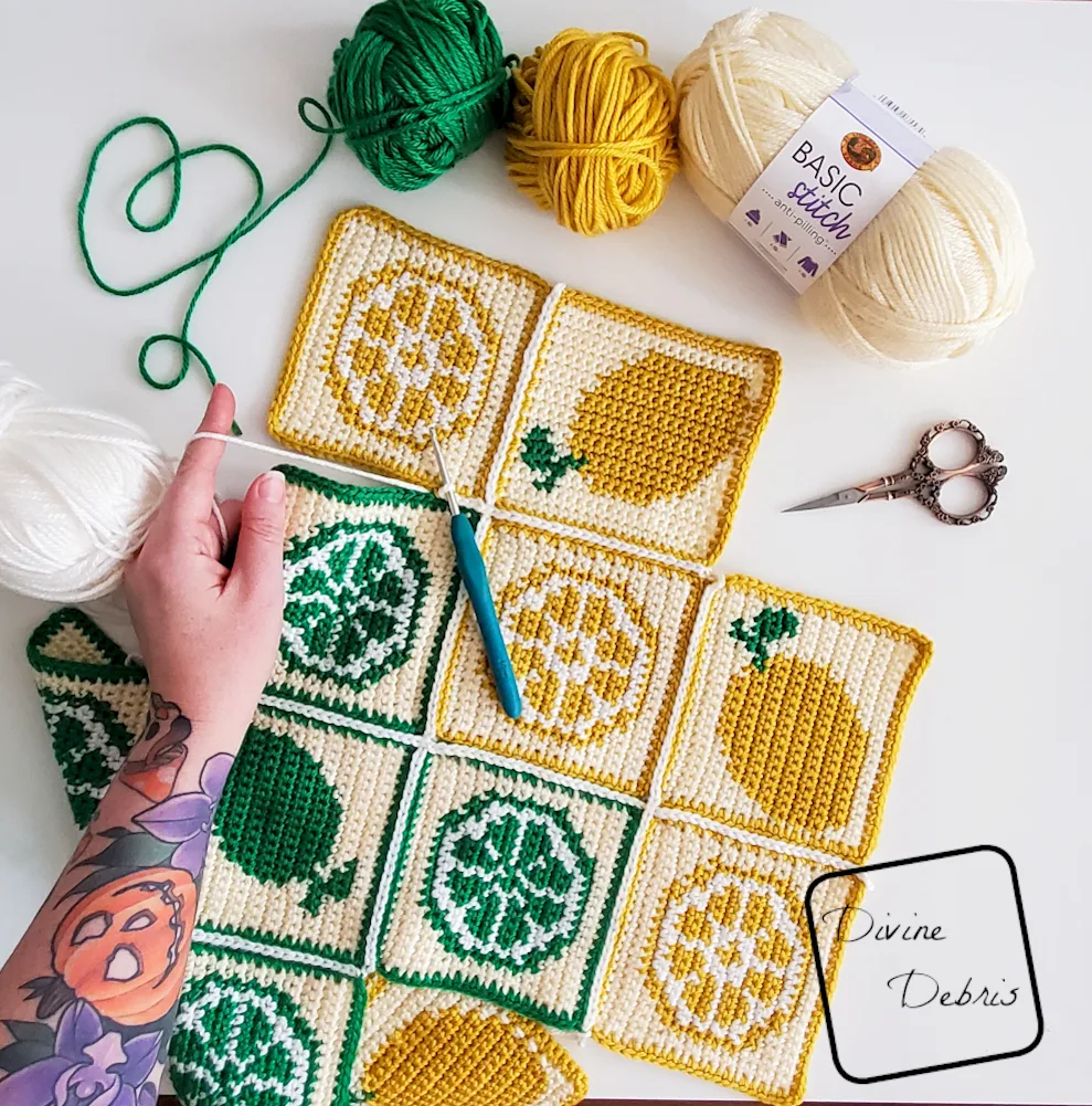 [Image description] Top down view of the Citrus Crush Bag still in squares being seamed, with a white woman's hand holding a strand of yarn.