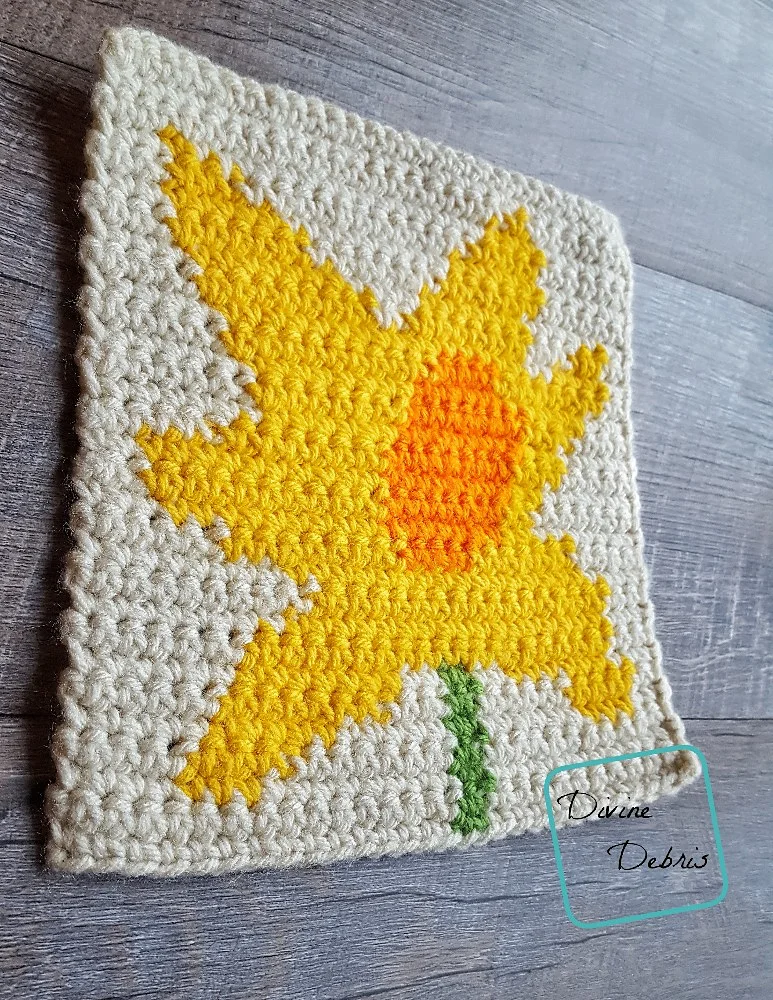 [Image description] a side angle of the 8" Tapestry Daffodil Afghan Square on a wood grain background