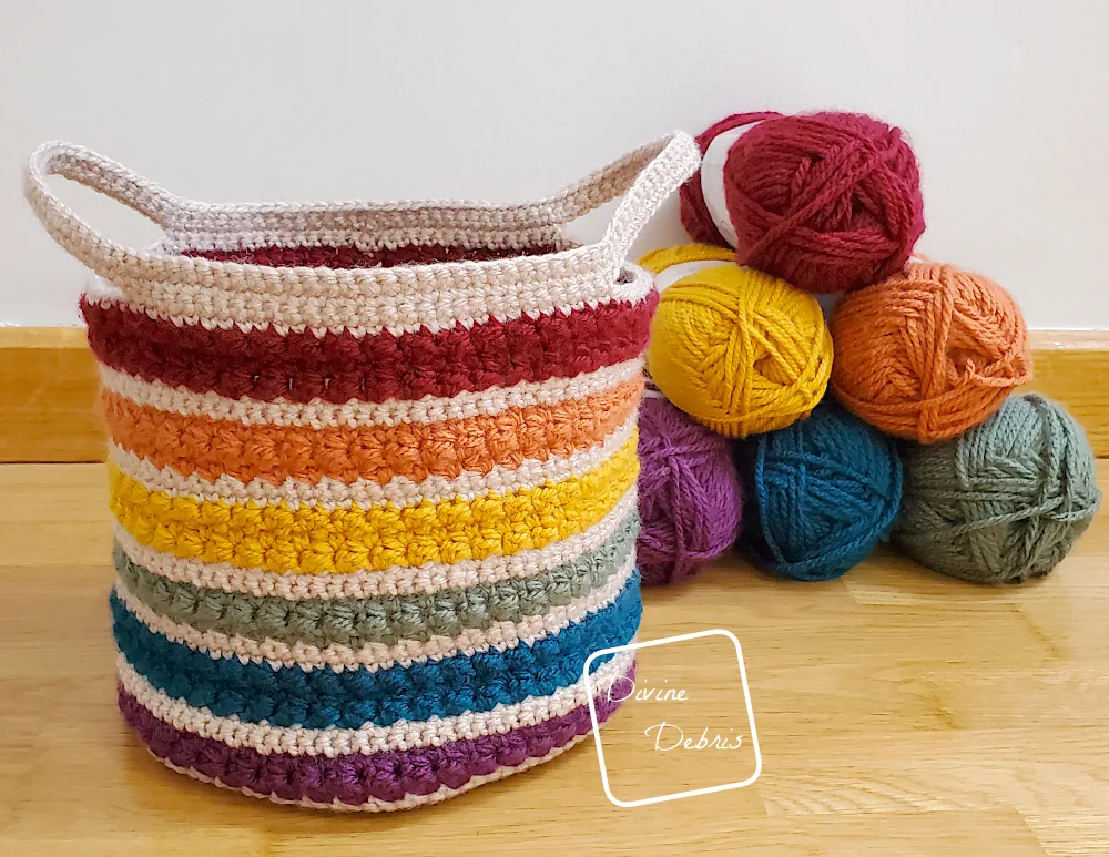[Image description] The Rainbow Layer Cake Basket sits on a wooden floor in front of a white wall and a pyramid stack of yarn skeins used to make the basket.