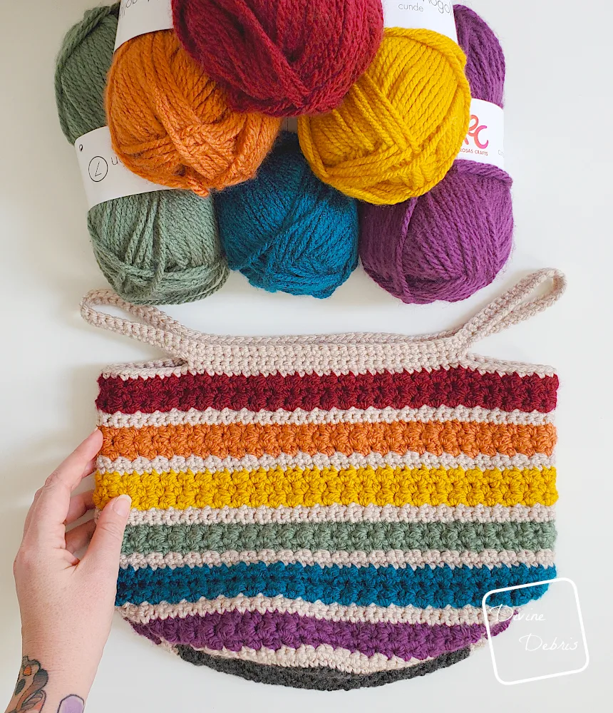 [Image description] Top down view of the Rainbow Layer Cake Basket laying flat on a white background, with skeins of yarn at the top of the photo and a white woman's hand holding the left side of the basket. 