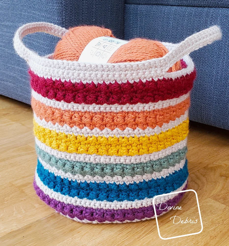 [Image description] Side photo of the Rainbow Layer Cake Basket sitting on a wooden floor next to a gray couch with a skein of yarn showing at the top of the basket.