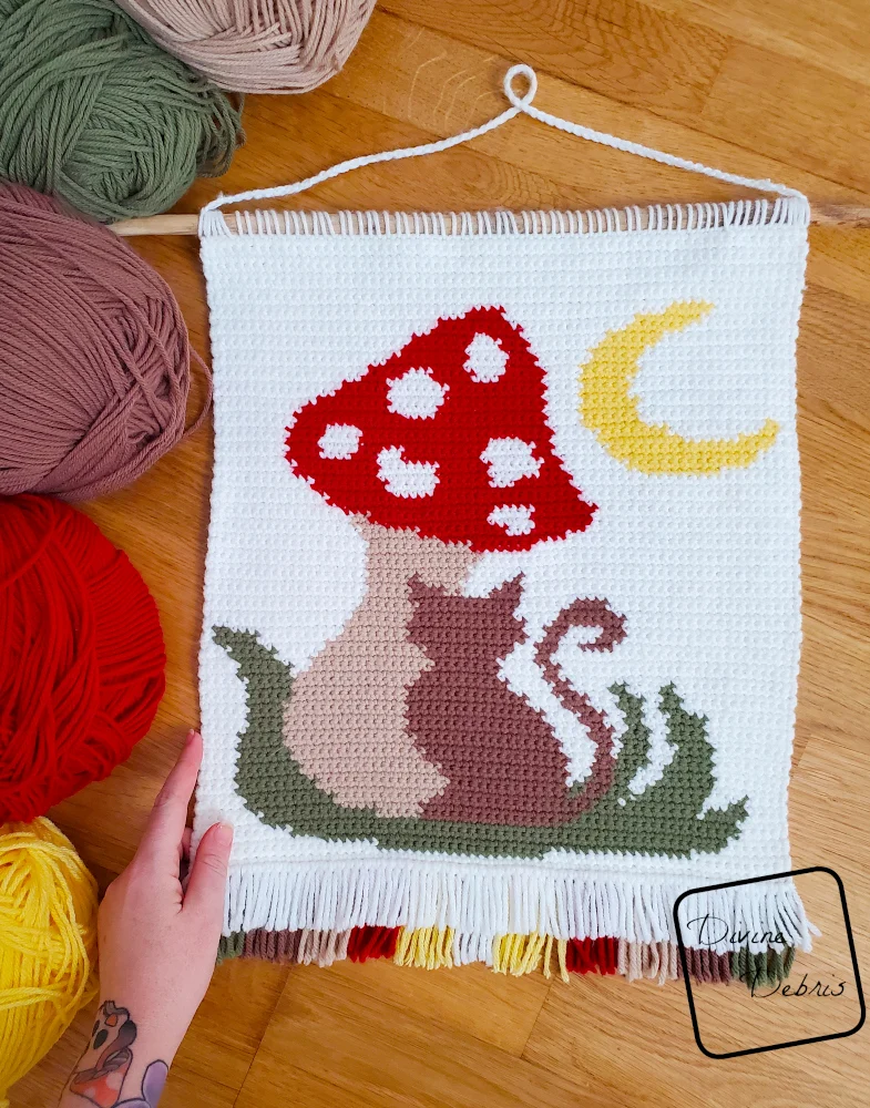 [Image description] Top down view of the Meow-Shroom Moon Wall Hanging laying on a wood grain background with skeins of yarn along the left side of the photo and a white woman's hand holding the wall hanging.