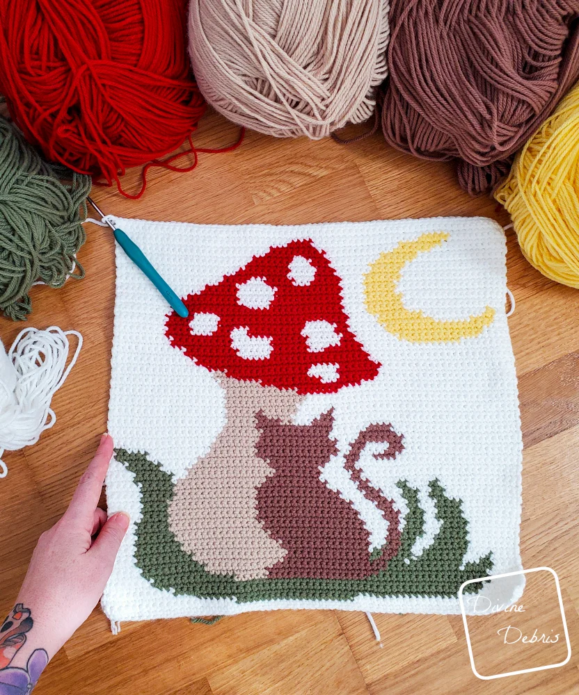 [Image description] top down view of an unfinished Meow-Shroom Moon Wall Hanging laying on a wood grain background with skeins of yarn along the top of the photo.