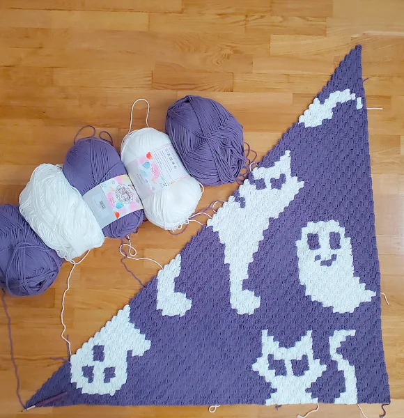 [Image description] Top down view of a half-finished A Ghoul's Best Friend Blanket with 5 skeins of yarn along the top of the photo.