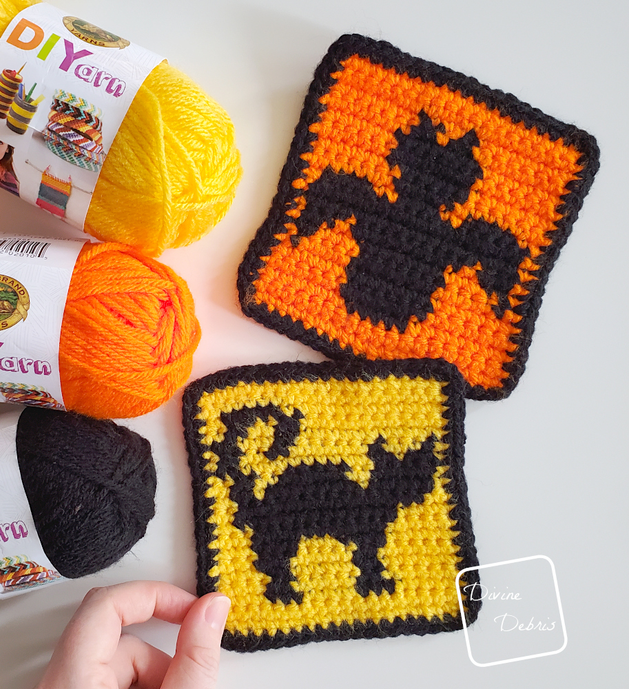 [Description} Top down view of the Cat and Bat Halloween Squares on a white background, with skeins of yarn along the left side of the photo, and a white woman's hand holding the cat square.