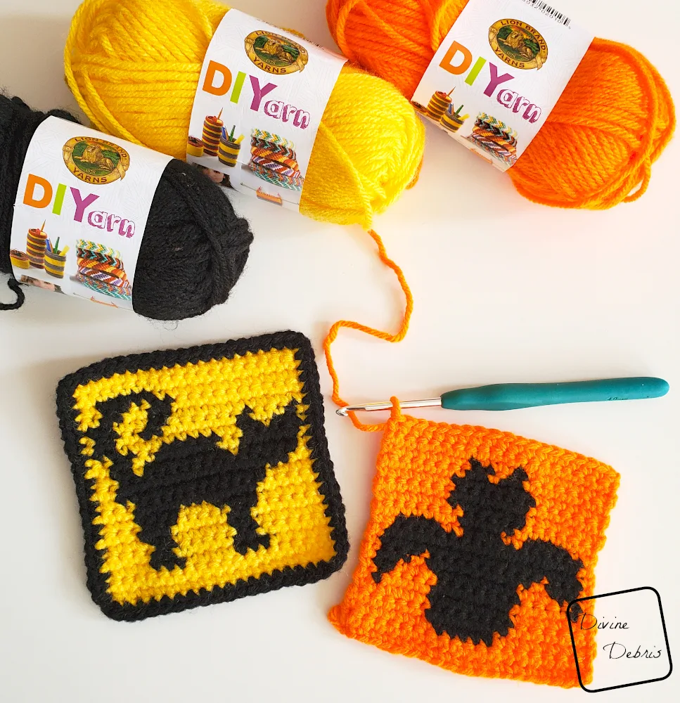 [Image description] Top down view of the Cat and Bat Halloween Squares, unfinished, on a white background with a crochet hook attached to the Bat square and skeins of yarn along the top of the photo.