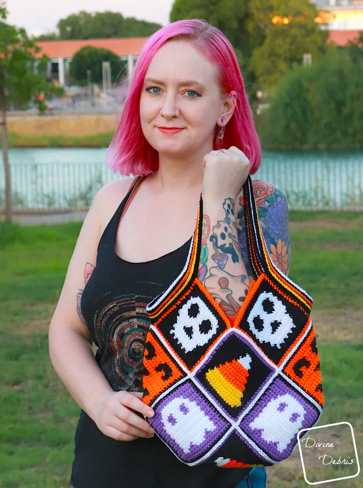 [Image description] A white woman with pink hair stands on grass holding the Patchwork Halloween Bag towards the camera.