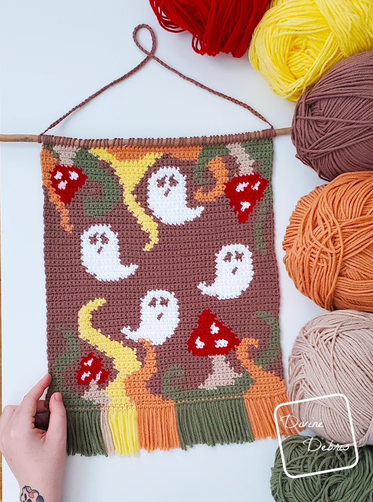 [Image description] Top down view of the Phantom Fungus Wall Hanging on a white background with skeins of yarn along the right side and a white woman's hand holding the bottom left-hand corner.