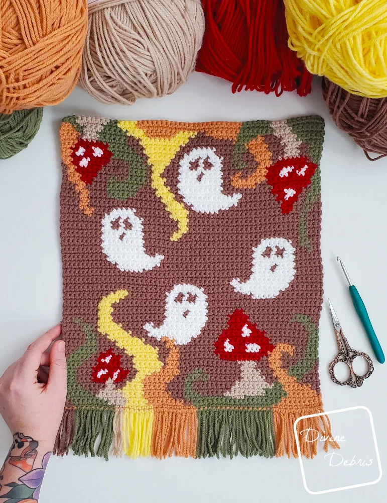 A unfinished Phantom Fungus Wall Hanging lays on a white surface with skeins of yarn along the top of the frame and a white woman's hand holds the bottom left-hand corner. 