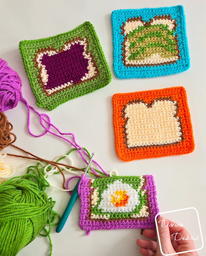 [Image description] The Avocado Toast, the Bread Square, the Jam Toast, and a half finished Avocado Egg Square with a white woman's hand holding the corner, are laying on a white background with skeins of yarn along the left side of the frame.