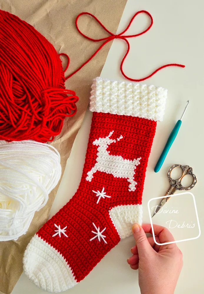 [Image description] A top down look at the Twinkle Toes Reindeer Stocking on a white and tan background with skeins of yarn along the top left and a white woman's hand holding the bottom right corner.