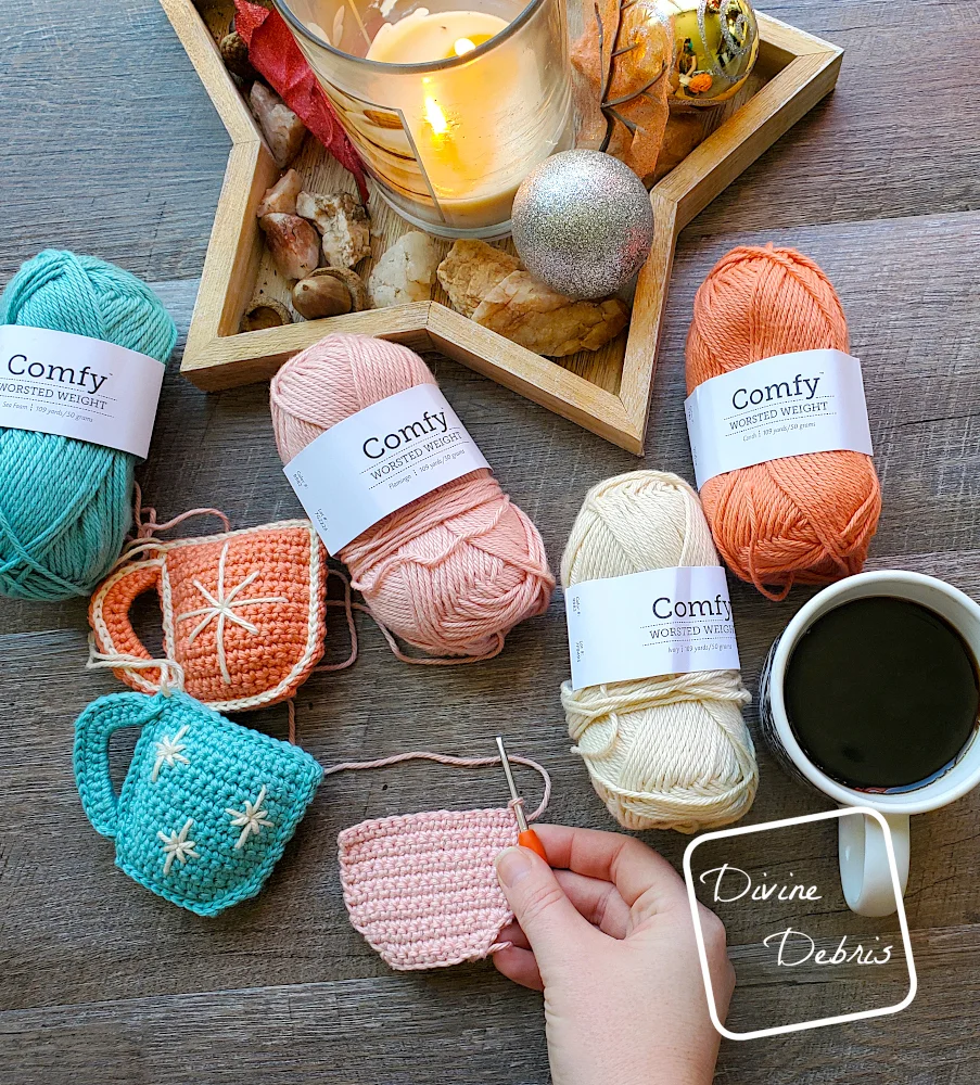 [Image description] Top down view of a white woman's hand holding an in progress Cute Cup Ornament, surrounded by skeins of yarn and finished Cute Cup Ornaments on a fake wood background.