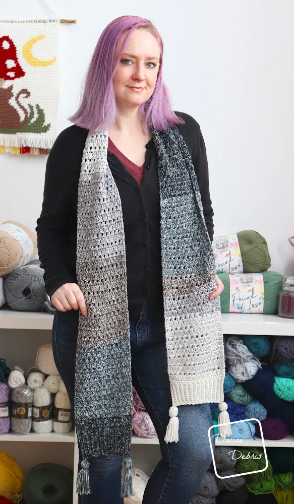 A white woman with purple hair stands in front of a white wall and shelves of yarn showing off the Blair Scarf hung long her neck.