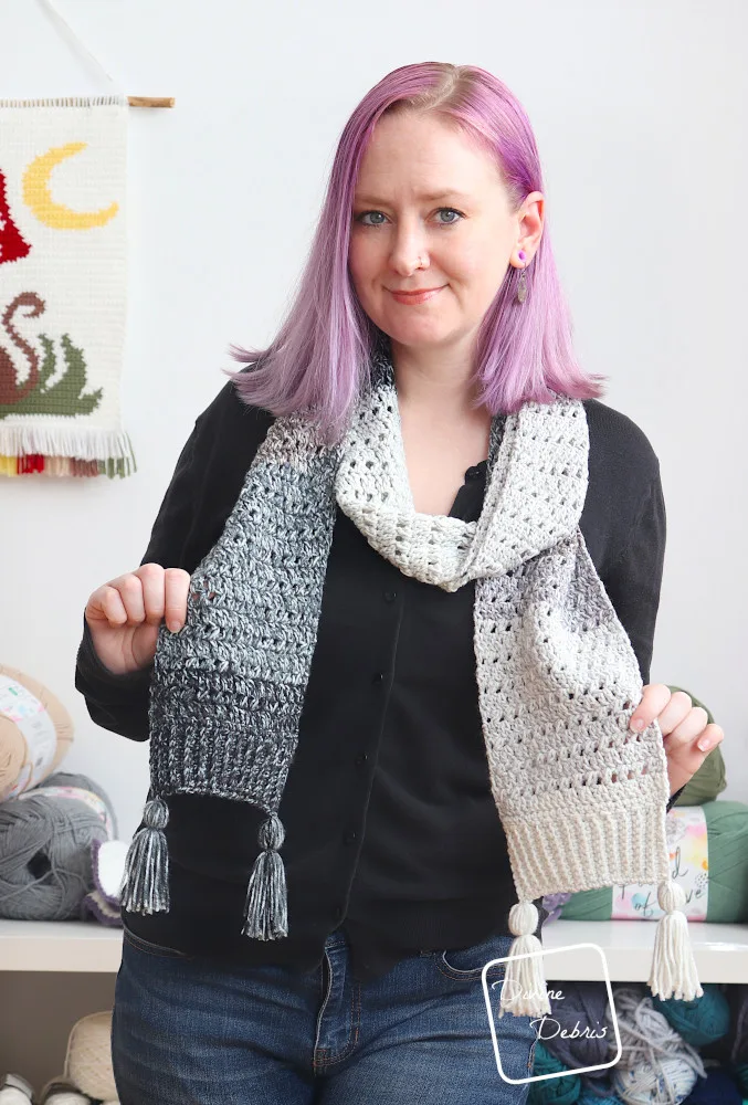 A white woman with purple hair stands in front of a white wall showing off the Blair Scarf wrapped around her neck, while holding up the ends to the camera.