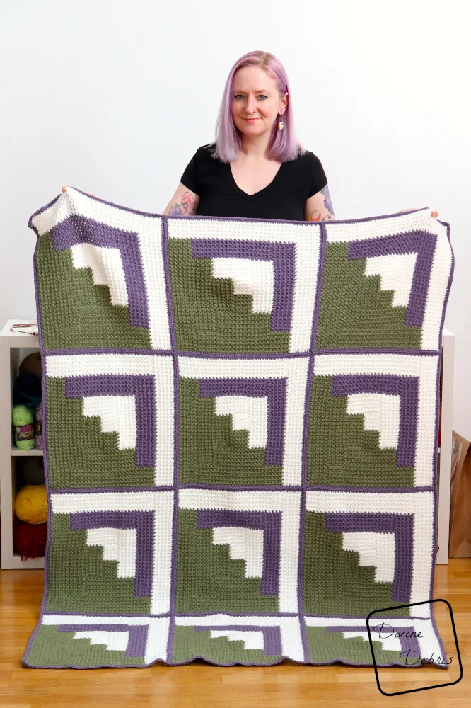 [Image description] A white woman with purple hair stands against a white background holding the Mountain Log Cabin Blanket towards the camera.