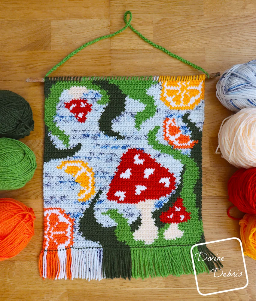 [Image description] Fruit of the 'Shroom Wall Hanging surrounded by skeins of yarn on both sides on a wood grain background.