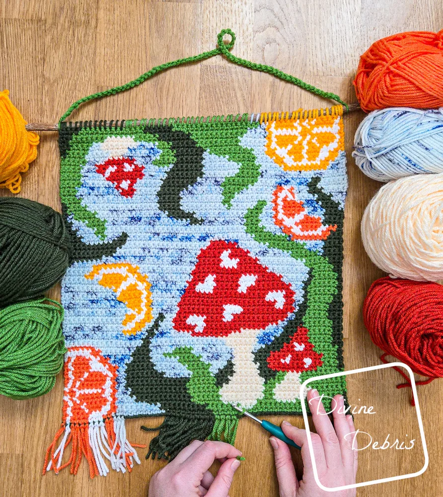 [Image description] Fruit of the 'Shroom Wall Hanging surrounded by skeins of yarn on both sides on a wood grain background and a white woman's hands adding fringe to the bottom.