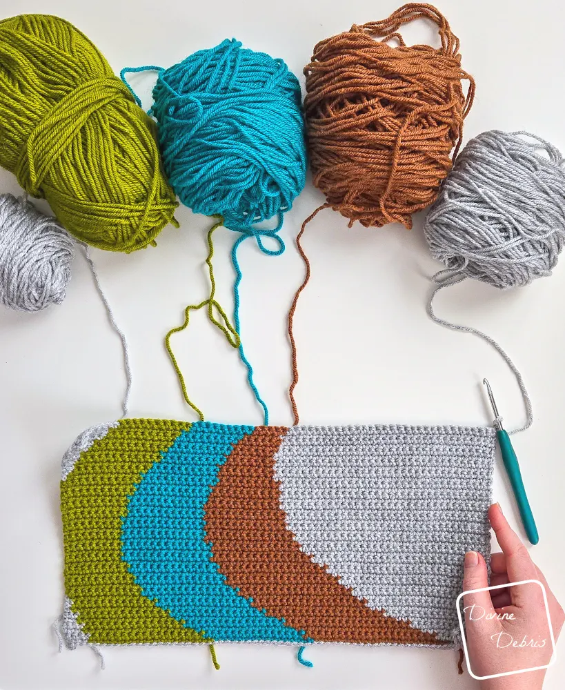 [Image description] Top down view of an unfinished Color Swirl Wall Hanging with a white woman's hand holding the bottom right and skeins of yarn along the top.