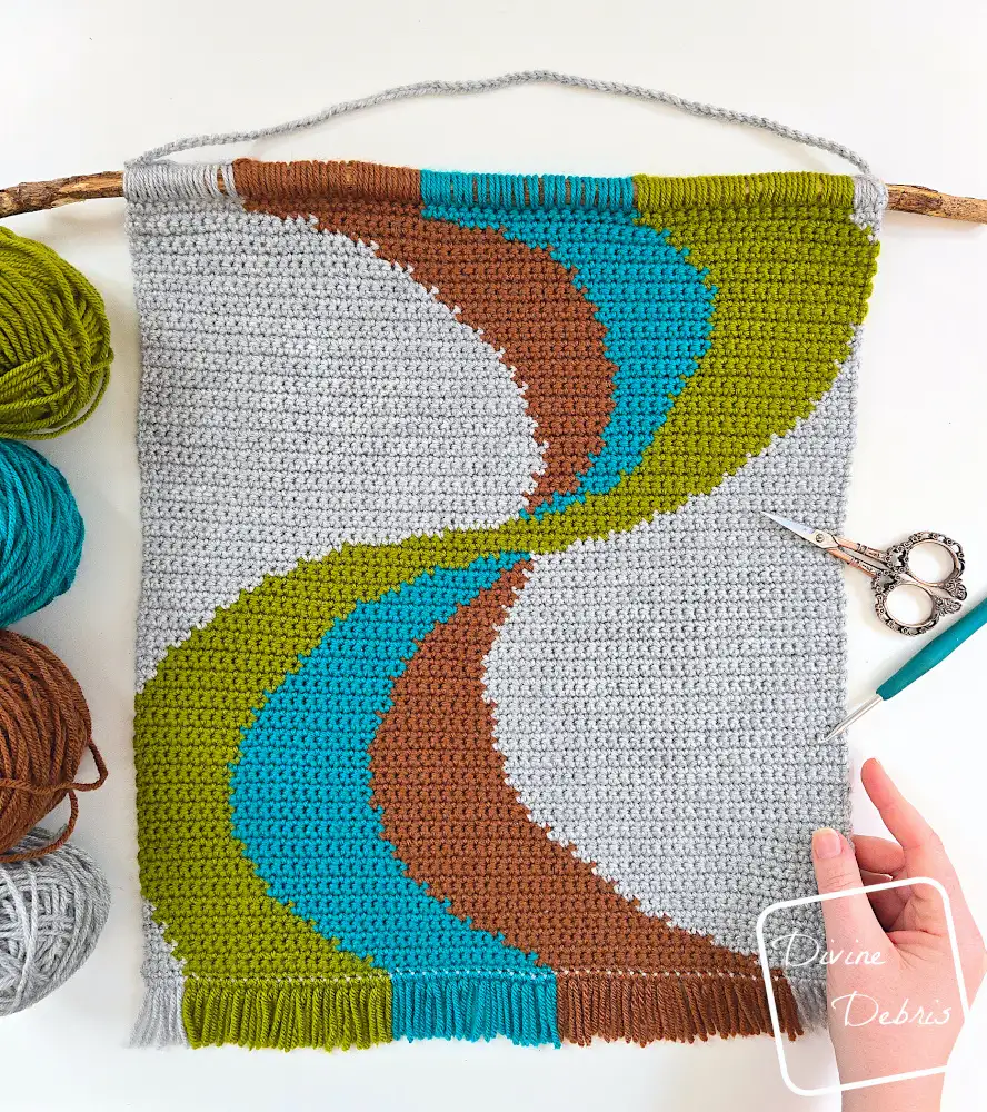 [Image description] Top down view of the Color Swirl Wall Hanging on a white background with a white woman's hand holding the bottom right-hand corner and skeins of yarn along the left side of the photo.