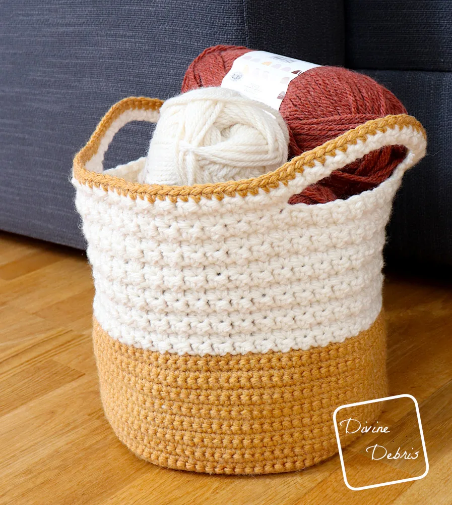 [Image description] The Trudy Two-Tone Basket shown sitting on a wood grain floor against a gray couch.