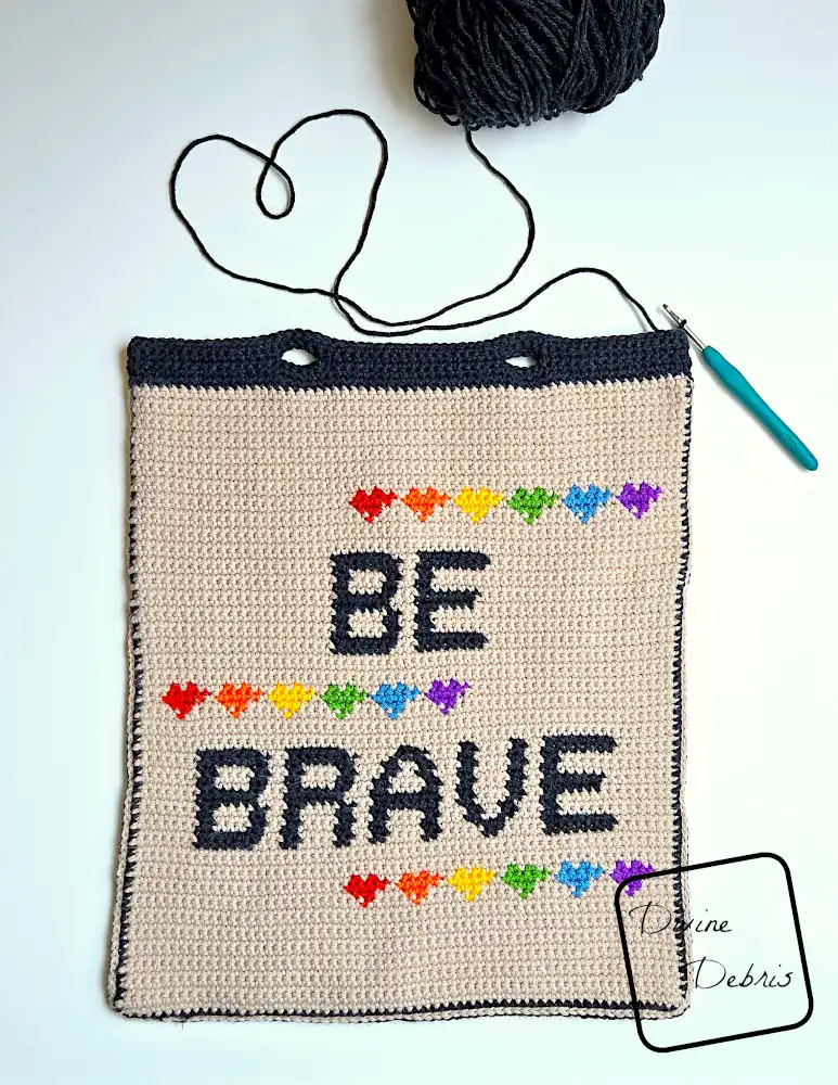 [Image description] The unfinished Be Brave Bag crochet pattern lays flat on a white background, with a skein of yarn on the top, a tail of yarn creating a heart, and a crochet hook attached.