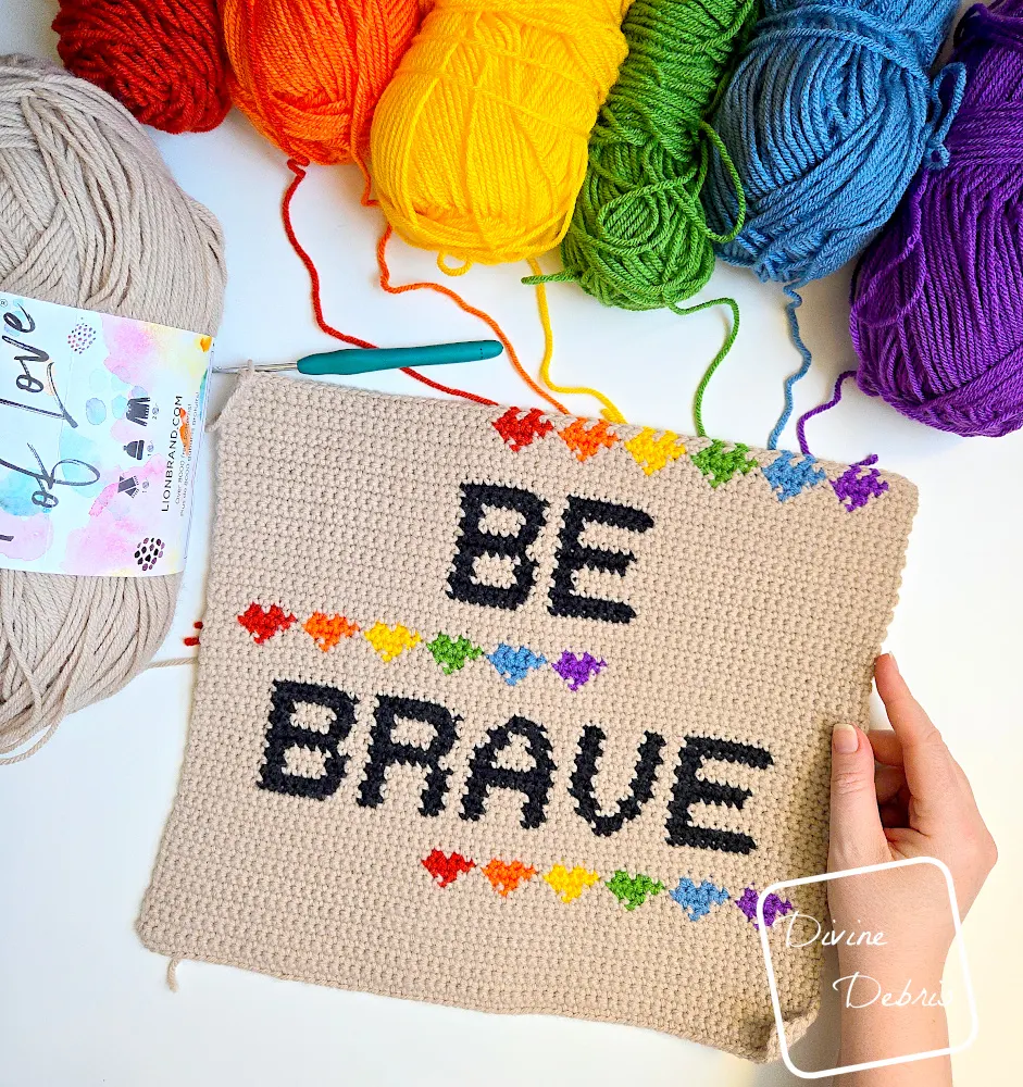 [Image description] Top down look at a unfinished version of the Be Brave Poster with a white woman's hand holding the right side of the design with rainbow colors of skeins of yarn along the top of the photo.