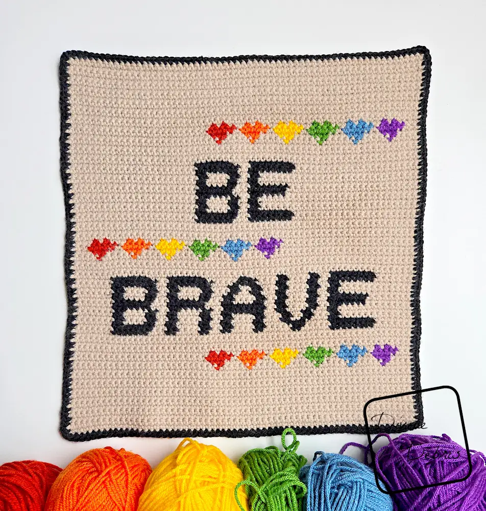 [Image description] Top down view of the Be Brave Poster on a white background, with a skeins of yarn along the bottom of the photo arranged in a rainbow.