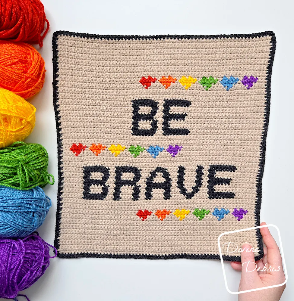 [Image description] Top down view of the Be Brave Poster on a white background, with a skeins of yarn along the left side of the photo arranged in a rainbow and a white woman's hand holding the bottom right-hand corner of the poster.