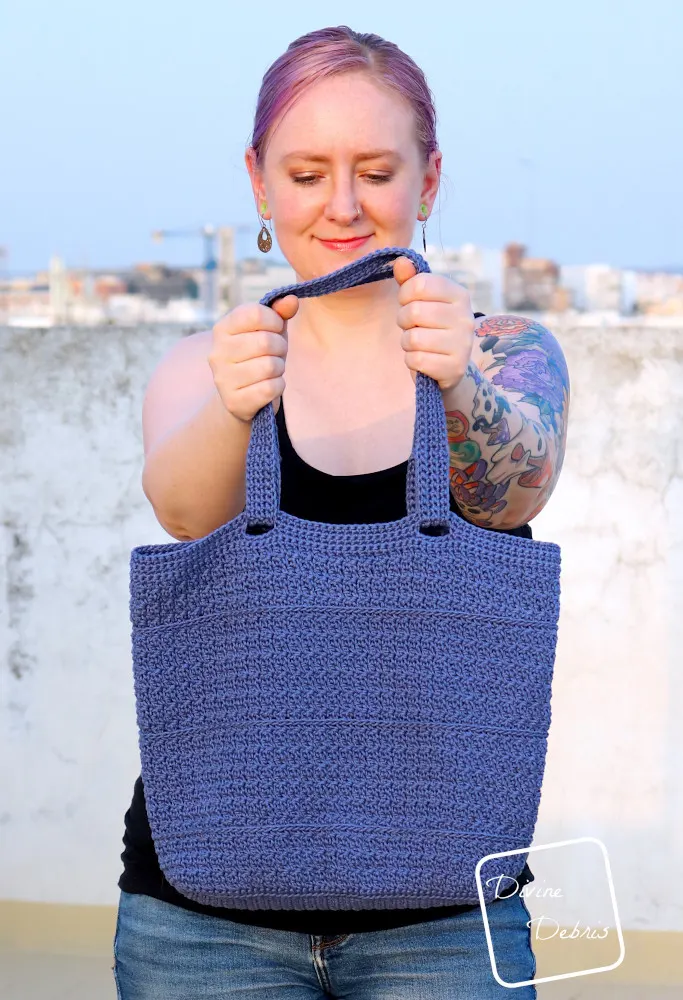 [Image description] A white woman with purple hair stands in front of a city view with the Kieran Bag positioned straight out toward the viewer.
