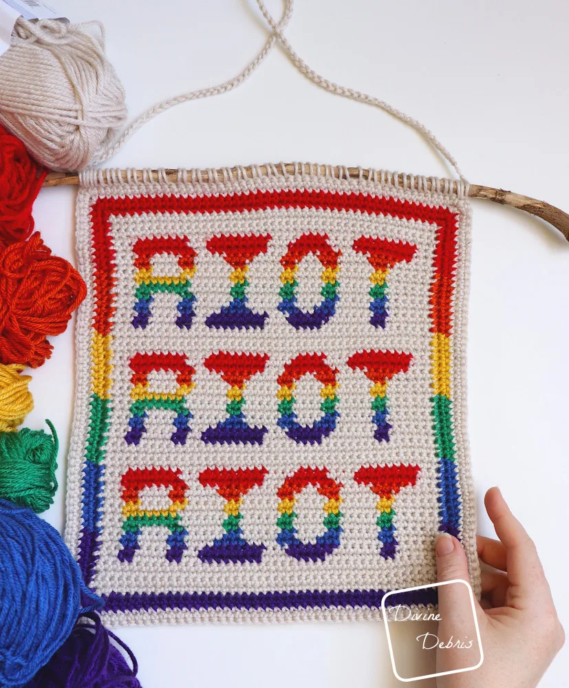 [Image description] Top down look at a Riot Wall Hanging crochet pattern laying on a white background with skeins of yarn along the left side of the photo and a white woman's hand holding the bottom right-hand corner.