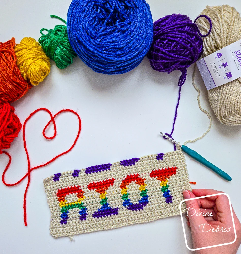 [Image description] Top down look at an unfinished Riot Wall Hanging crochet design - with skeins of yarn along the top of the photo and a white woman's hand holding the bottom right-hand corner of the wall hanging.