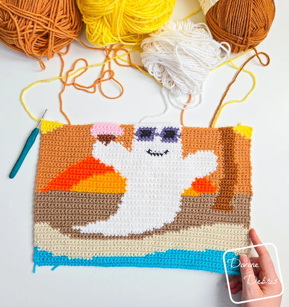 [Image description} An unfinished photo of the Beach Ghost Wall Hanging on a white background, with skeins of yarn along the top of the photo and a white woman's hand holding the bottom right-hand corner.