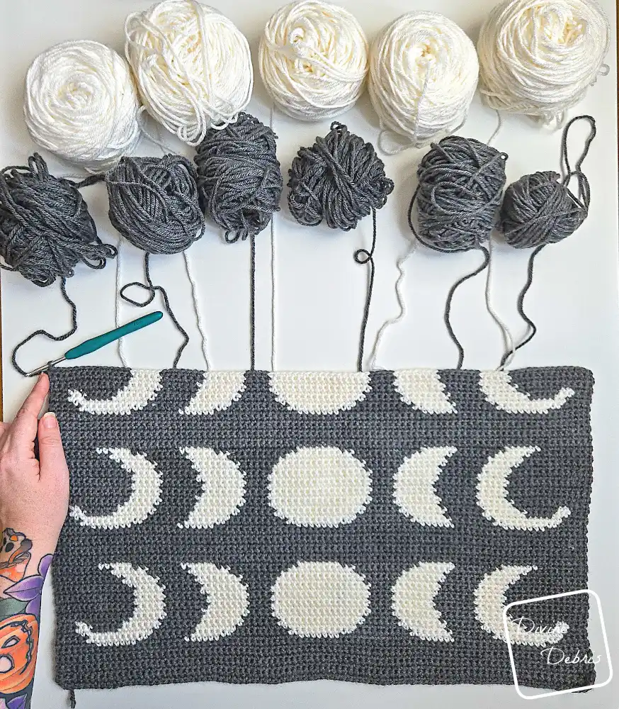[Image description] Top down look at an unfinished Moon Phase Wall Hanging crochet design - with skeins of yarn along the top of the photo and a white woman's hand holding the bottom left-hand corner of the wall hanging.
