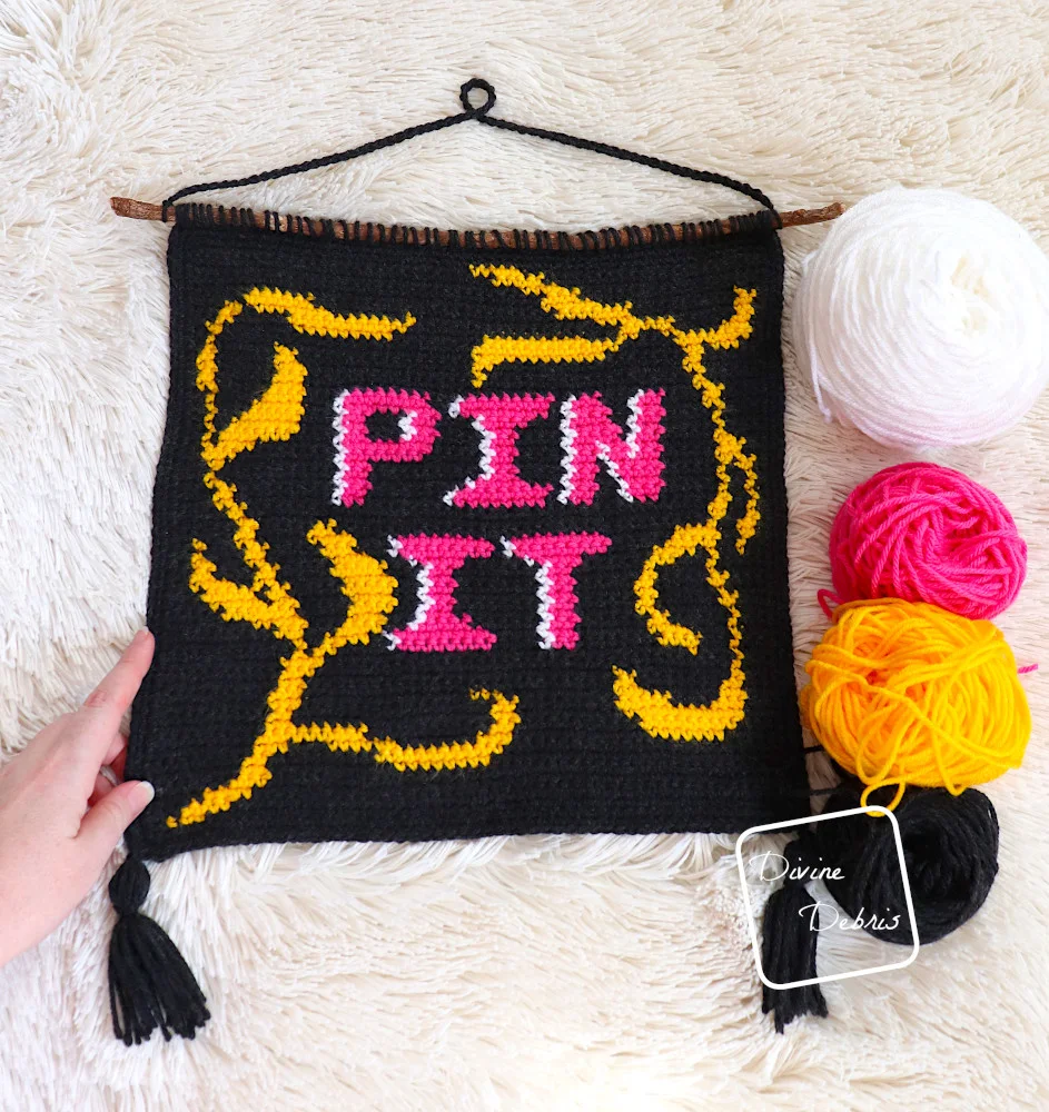 [Image description] The Pin It Wall Hanging lays on a fuzzy white blanket with skeins of yarn along the right side and a white woman's hand holds the left side bottom corner.