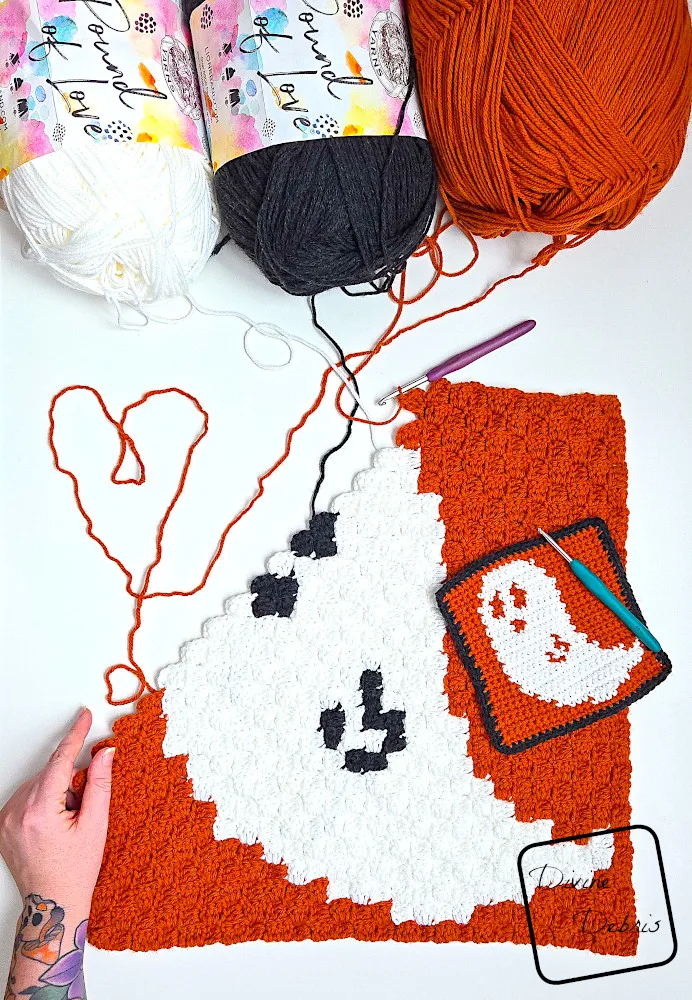 [Image description] top down view of an unfinished Gwendoline Ghost C2C Square with skeins of yarn along the top of the frame and a white woman's hand holding the bottom corner.