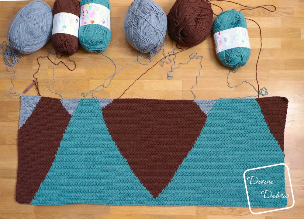[Image description] An unfinished Moon Rise Mountains Blanket lays flat on a wood grain floor with skeins of yarn along the top of the photo to illustrate how the yarn was still attached to the blanket while it was being made.