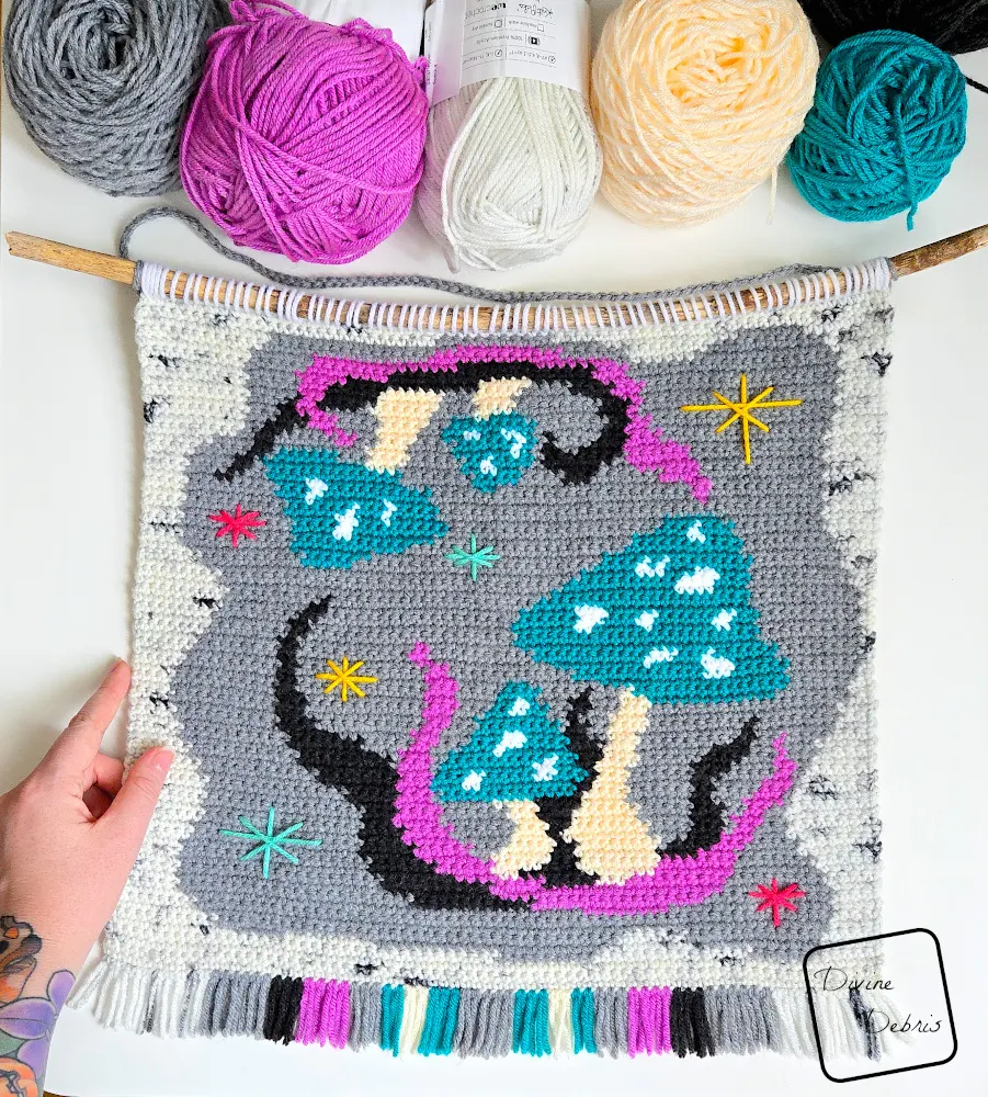 [Image description] Top down look at the Wild Mushrooms Wall Hanging on a white background with skeins of yarn along the top of the photo and a white woman's hand holding the bottom left corner.