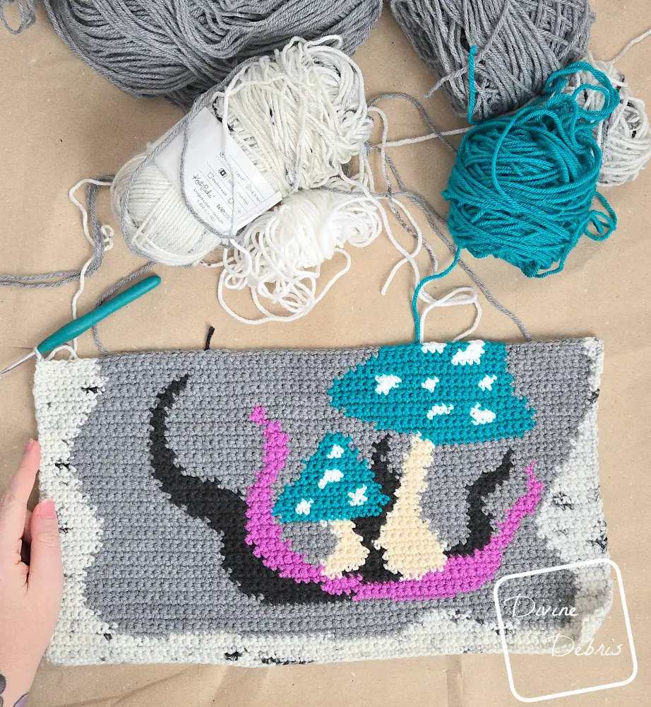 [Image description] Top down look at the Wild Mushrooms Wall Hanging in progress with skeins of yarn messily on the top of the photo and a white woman's hand holding the bottom left corner.