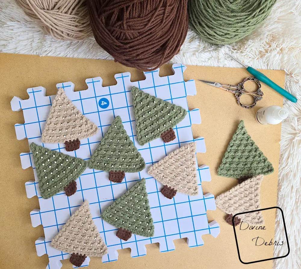[Image description] Top down look at a collection of trees for the Cute Tree Garland crochet pattern on a graph design blocking square and a tan board on a white blanket. 