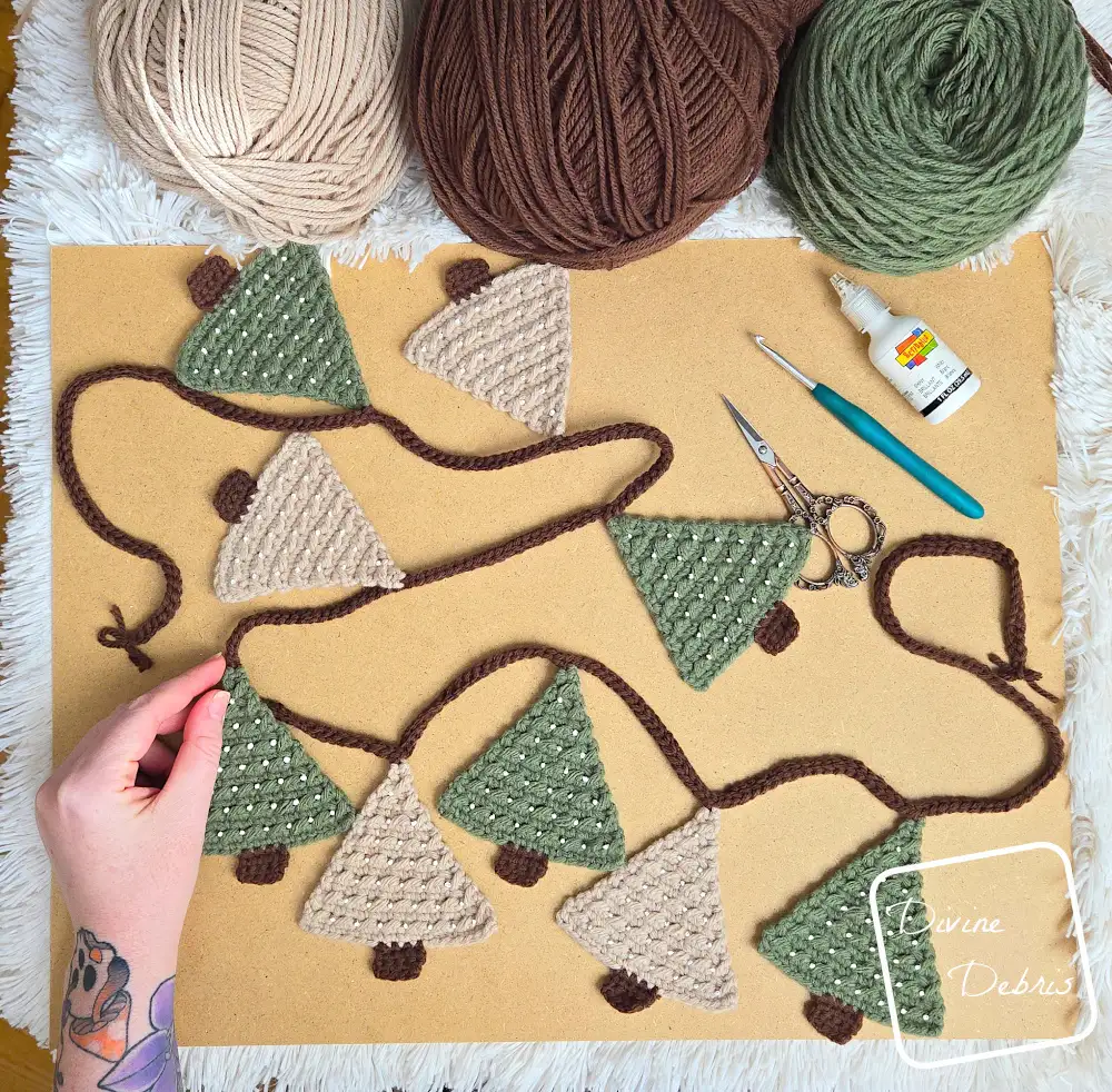 [Image description] Top down look of a white woman's hand holding one tree on the Cute Tree Garland crochet pattern, laying on a tan background with a white background behind that and skeins of yarn along the top of the photo.