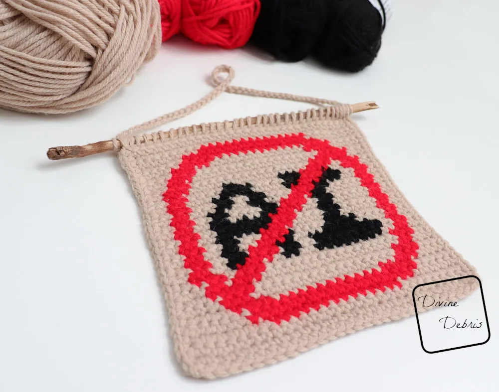 [Image description] A low angle photo of the Anti AI Wall Hanging on a white background with skeins of yarn along the top of the photo. 