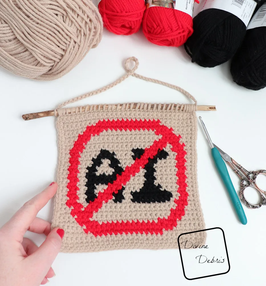 [Image description] Top down look at a finished Anti AI Wall Hanging laying on a white background with skeins of yarn along the top of the photo and a white woman's hand holding the bottom left-hand corner. 
