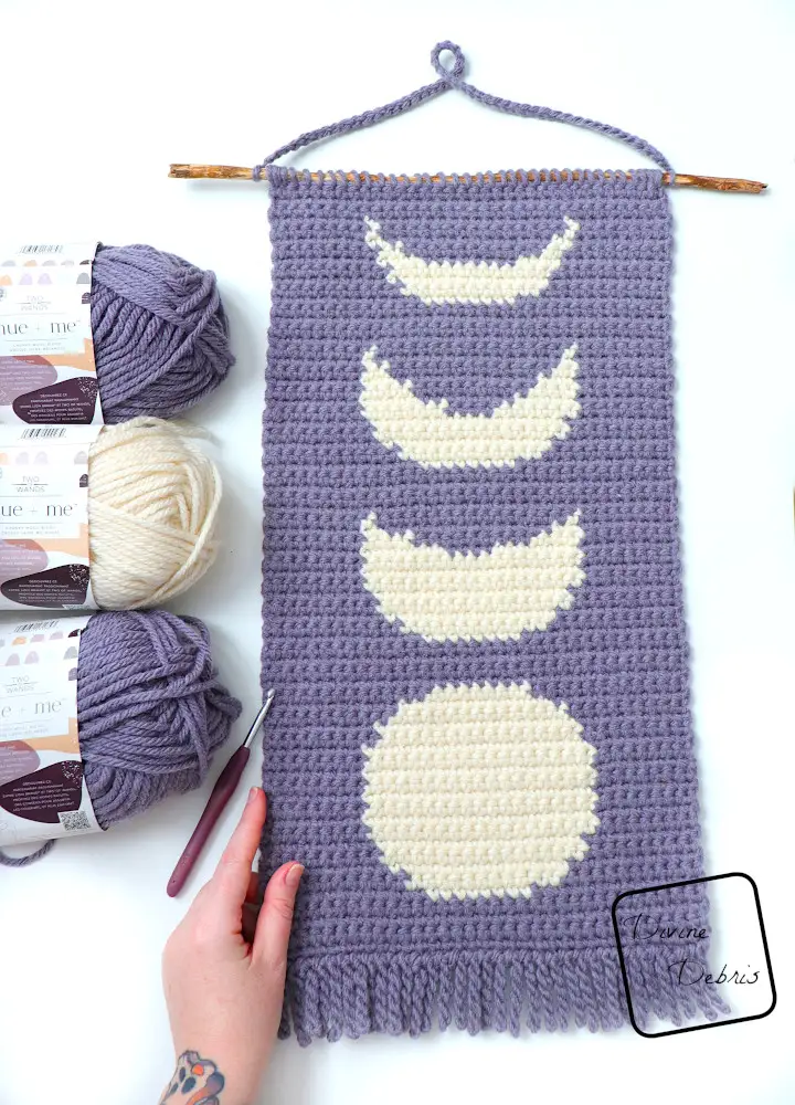 [Image description] The Simple Moon Phase Wall Hanging laying on a white background with a white woman's hand holding the bottom left hand corner.